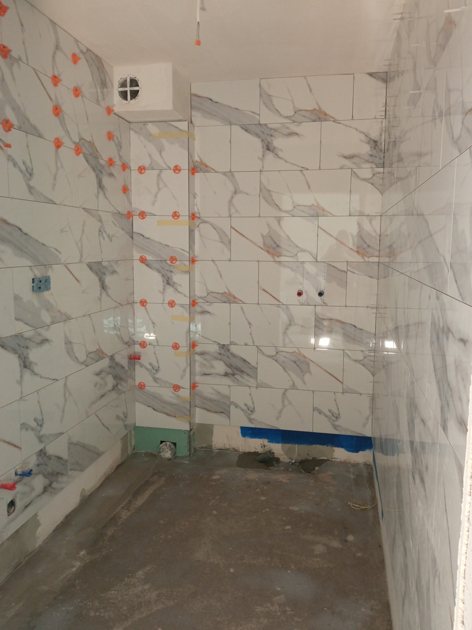 Montaż płytek imitujących marmur na ścianach łazienki, z użyciem systemów poziomowania płytek i taśmy malarskiej, widoczne instalacje wodne i wentylacja.