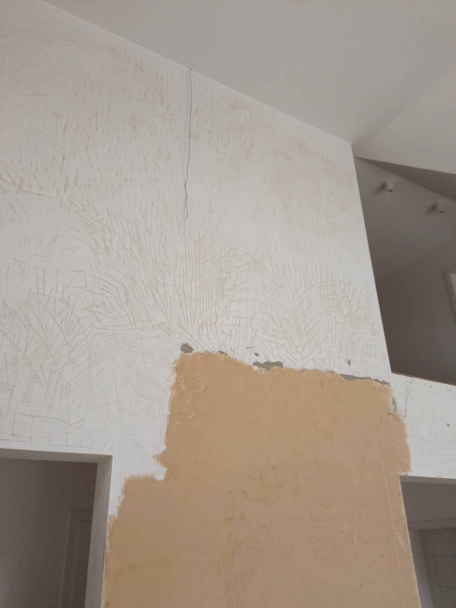 Przygotowanie ściany do malowania: zarysowana powierzchnia podłoża i fragment ściany z nałożoną warstwą podkładową w kolorze beżowym, widoczna szczelina w górnej części ściany.