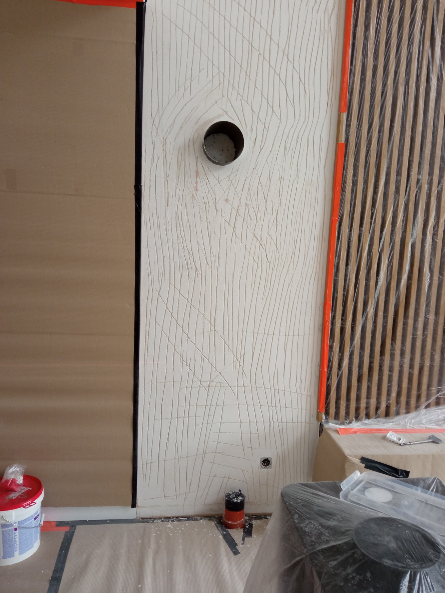 Ściana z dekoracyjną fakturą imitującą rysunek drewna, z otworem na rurę, w trakcie prac wykończeniowych, zabezpieczona folią i taśmą malarską.
