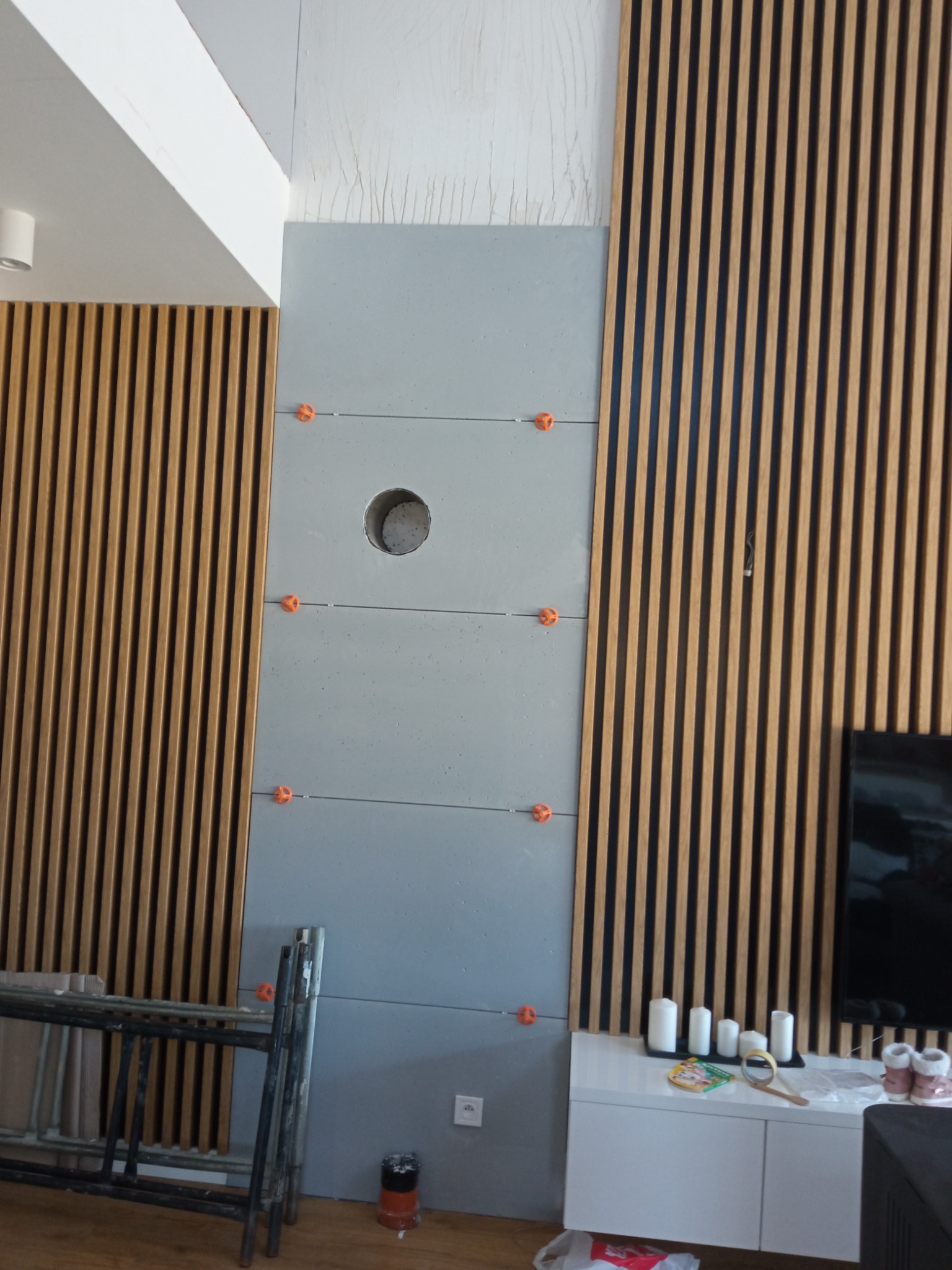 Montaż paneli ściennych: szare płyty z dystansami krzyżykowymi i drewniane lamele. Widoczne narzędzia i drabina.