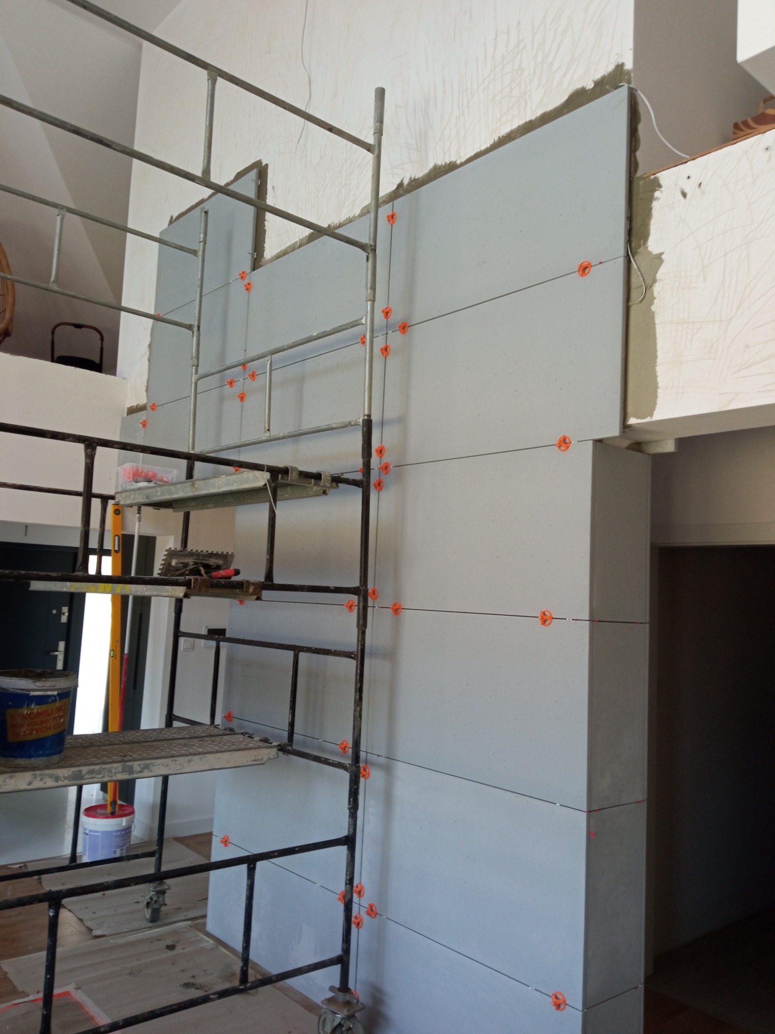 Montaż szarych płytek na ścianie z użyciem poziomych sznurków i pomarańczowych krzyżyków dystansowych, widoczne rusztowanie z narzędziami, etap prac glazurniczych.