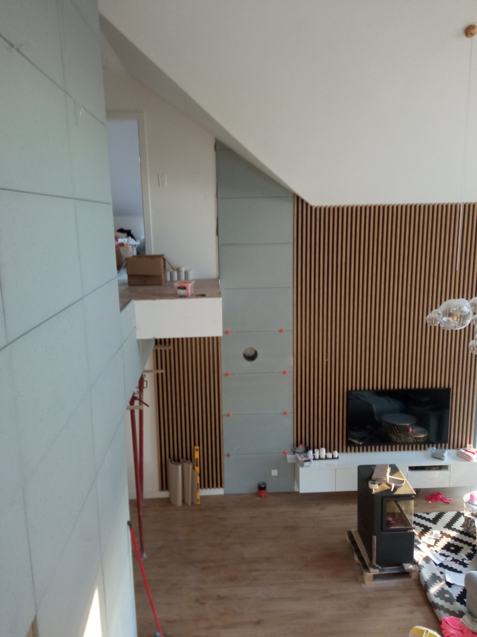 Wysoki salon w trakcie wykańczania, widoczne panele ścienne z drewna, częściowo obłożona ściana płytami, poziomica, kartony, oraz kominek na paletach przygotowany do montażu.
