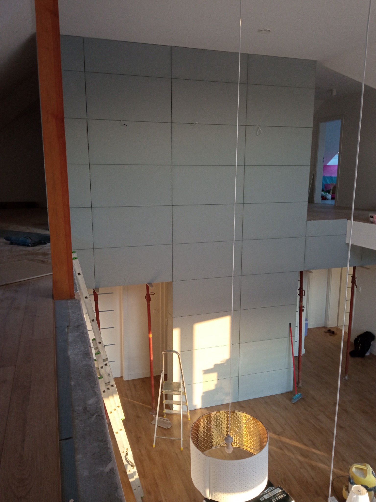 Wysoki pokój w trakcie prac wykończeniowych, widoczne szare panele na ścianie, drabiny, drewniana podłoga, lampa wisząca i podpory stropowe.