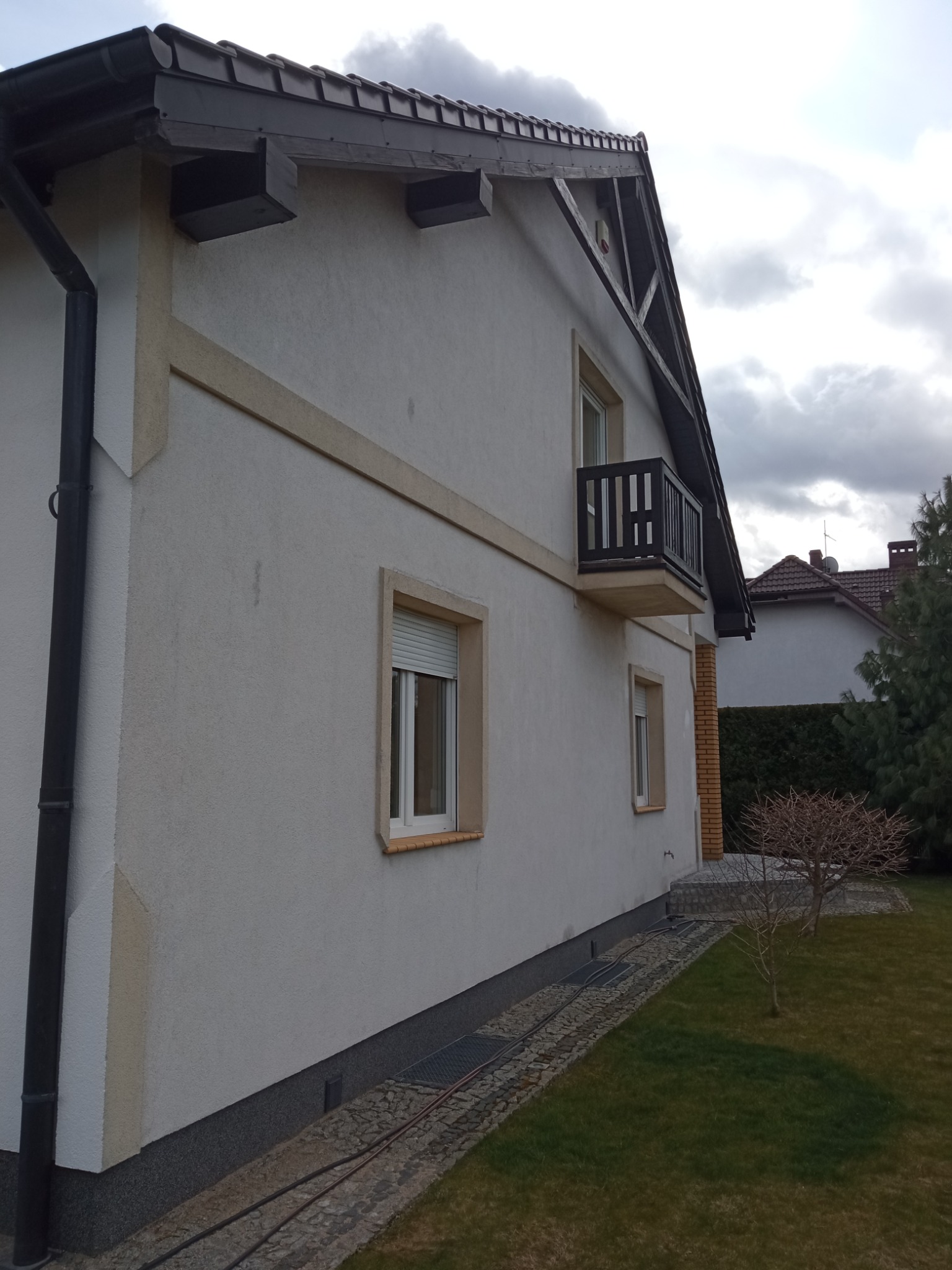 Boczny widok domu z białym tynkiem, ciemnobrązowym dachem i balkonem z czarną balustradą, widoczne okna z roletami i fragment trawnika.