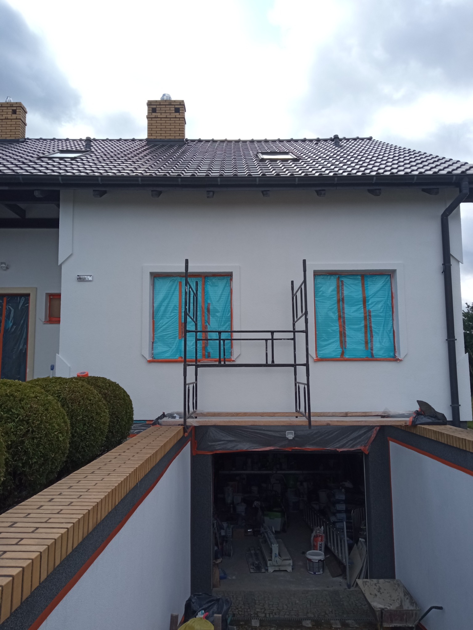 Przygotowania do malowania elewacji domu: okna zabezpieczone folią, rusztowanie ustawione, widoczny wjazd do garażu z narzędziami i materiałami.