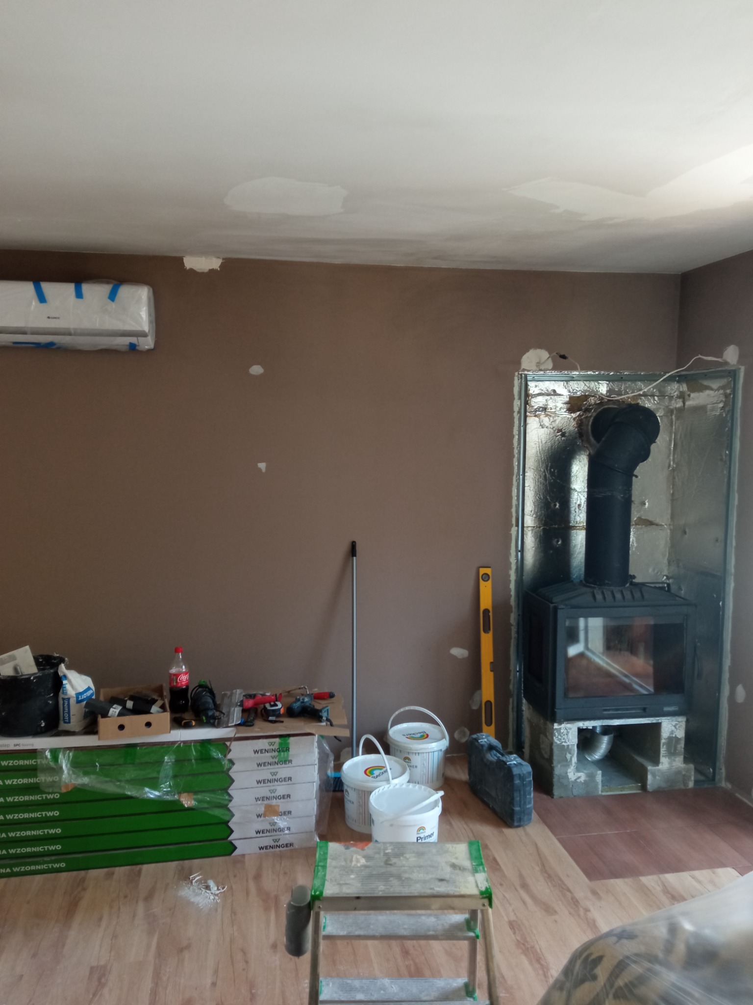 Wnętrze w Gorzowie Wielkopolskim w trakcie prac remontowych: ściana pomalowana na brązowo z widocznymi łatami, zabudowa kominka w trakcie tworzenia, panele podłogowe w opakowaniach, drabina...