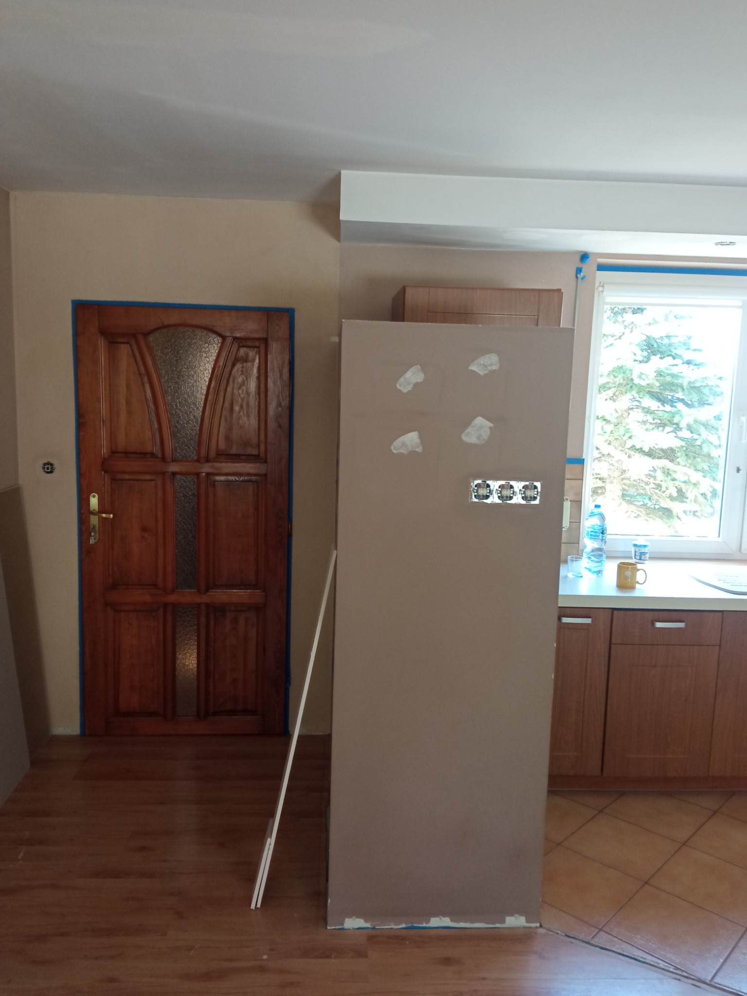 Remont kuchni w trakcie: ściana przygotowana do malowania z widocznymi punktami elektrycznymi, zabezpieczona taśmą brązowa drewniana stolarka okienna i drzwiowa, podłoga częściowo wyłożona...