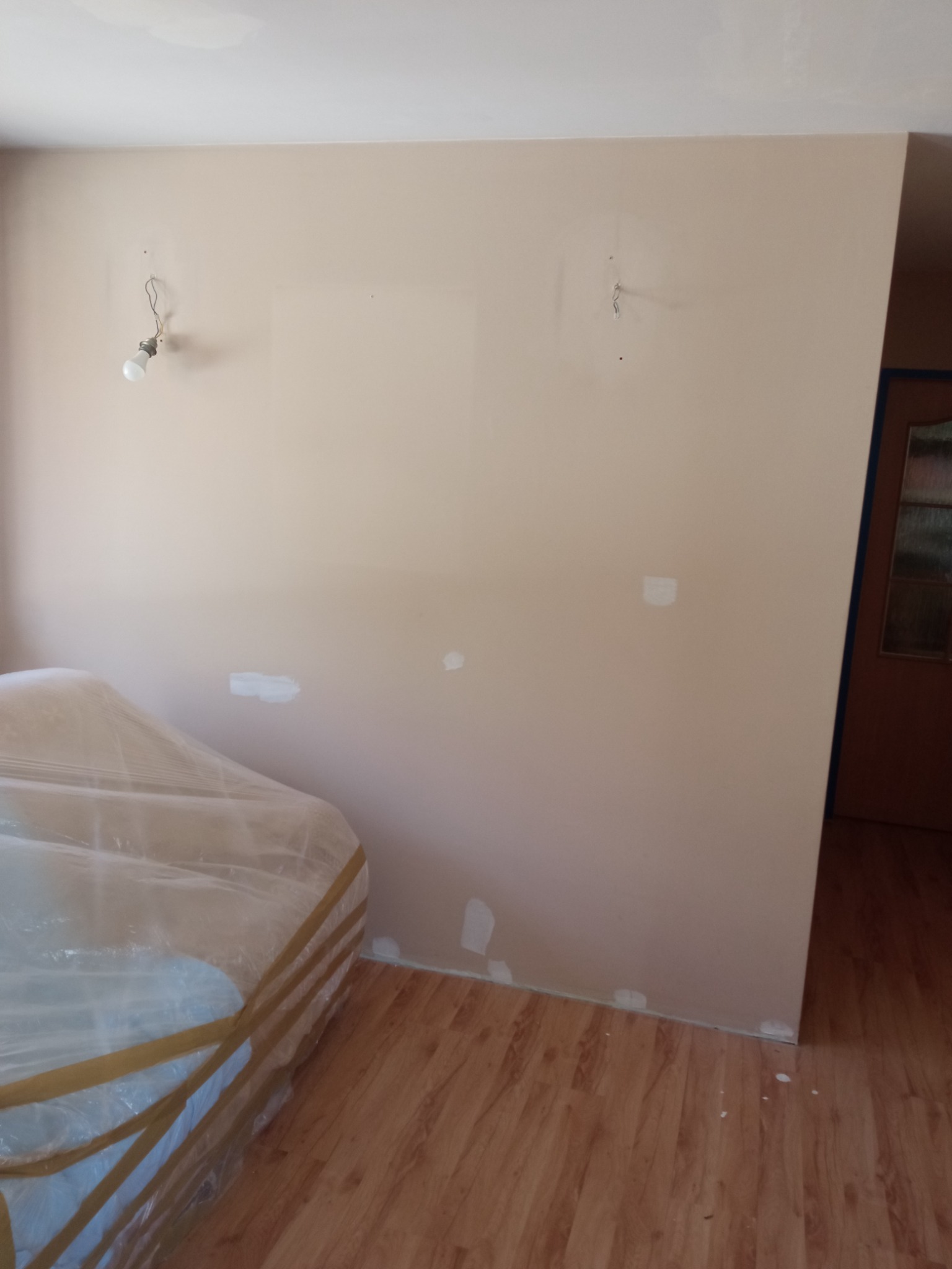 Przygotowana do malowania ściana w odcieniach beżu z widocznymi śladami po szpachlowaniu, zabezpieczone meble folią, surowa żarówka zwisająca z kabla, parkiet na podłodze.
