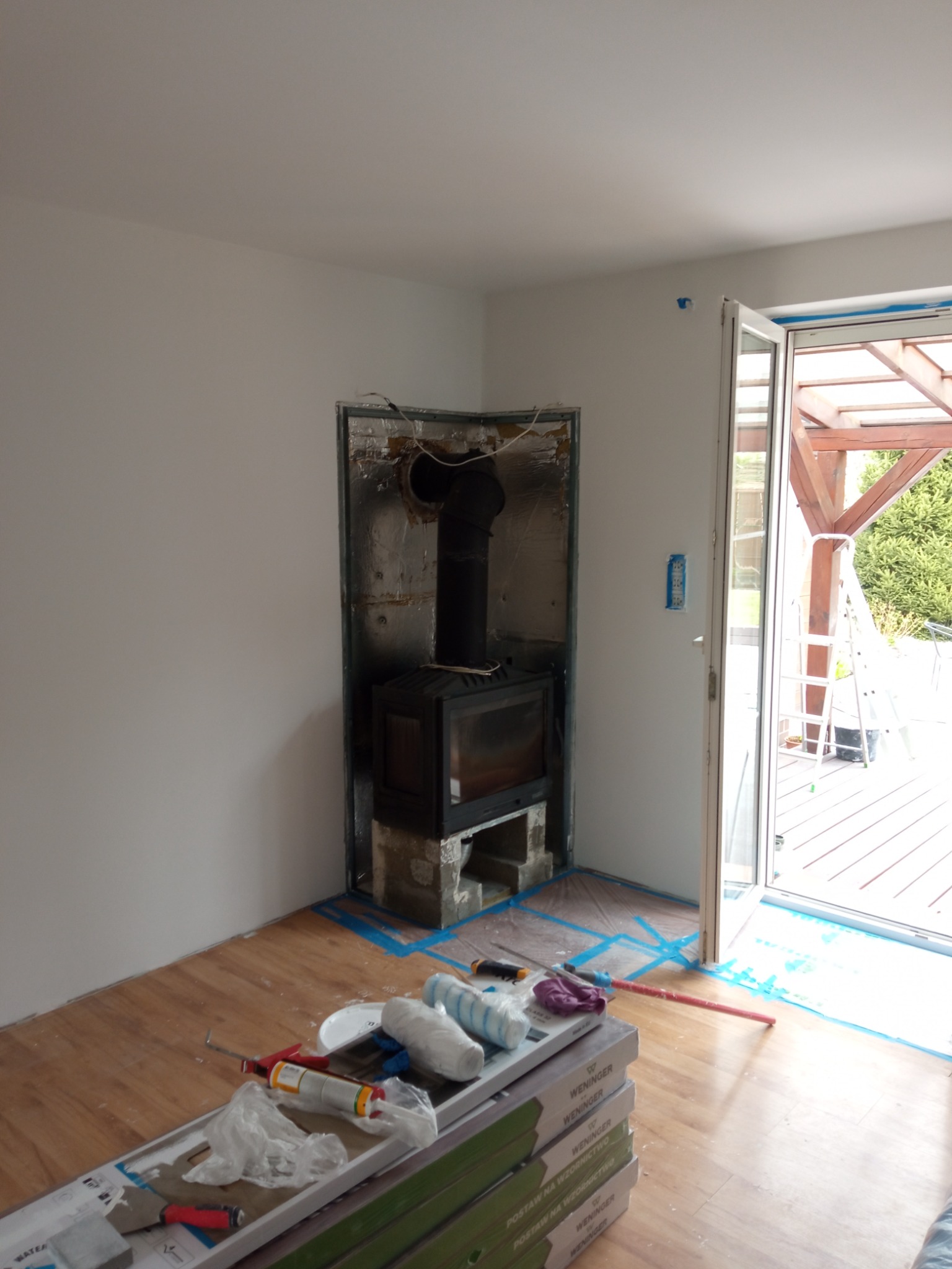 Przygotowania do obudowy kominka w surowym stanie, widoczne materiały budowlane, taśmy zabezpieczające oraz otwarte drzwi balkonowe z widokiem na taras.