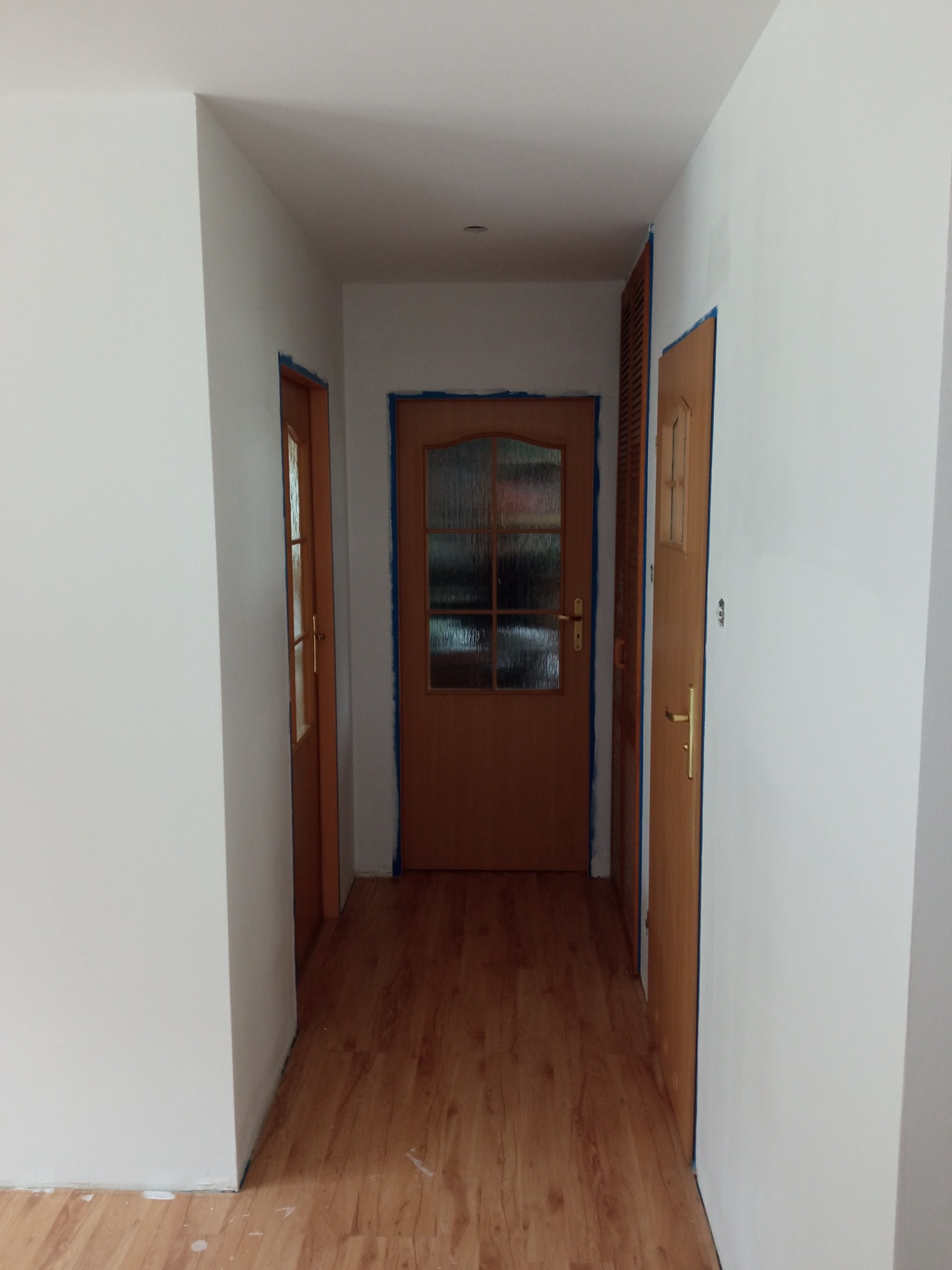 Korytarz w trakcie malowania: białe ściany, drewniana podłoga, drzwi zabezpieczone niebieską taśmą malarską, widoczne ślady farby na podłodze.