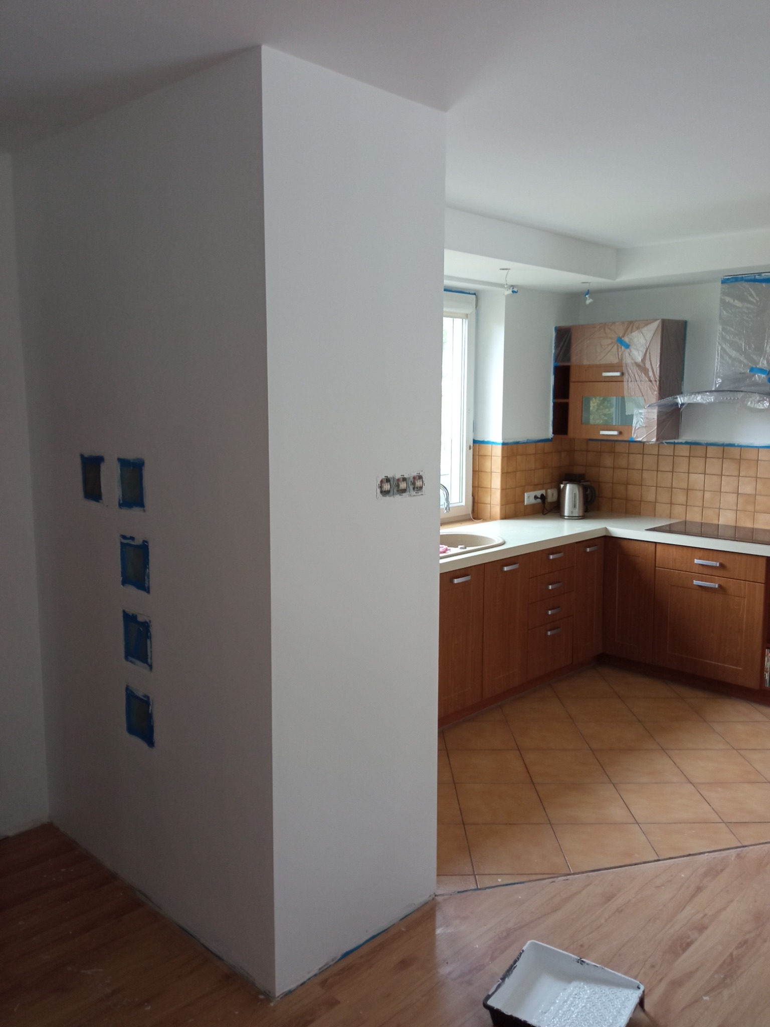 Świeżo pomalowana na biało ściana z zabezpieczonymi taśmą otworami na gniazdka elektryczne, widok na kuchnię z brązowymi meblami zabezpieczonymi folią malarską, pojemnik z farbą na podłodze.