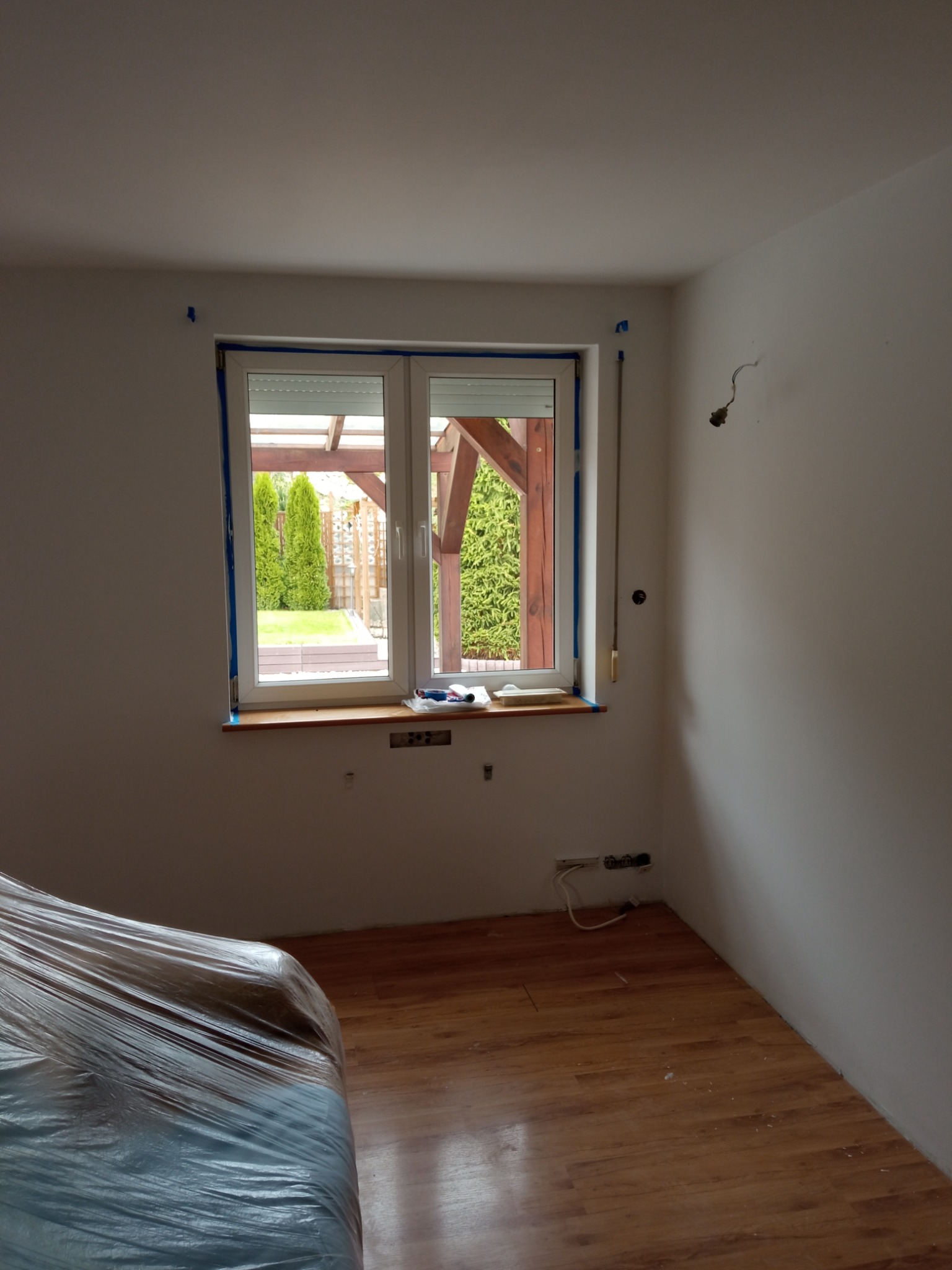 Pokój w trakcie przygotowania do malowania: okno zabezpieczone niebieską taśmą malarską, podłoga częściowo zakryta folią ochronną, widoczne gniazdka elektryczne i wystające przewody.