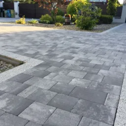 Ułożona szara kostka brukowa na podjeździe w Lipiu, z obrzeżem z jasnego kruszywa i elementami małej architektury ogrodowej w tle.