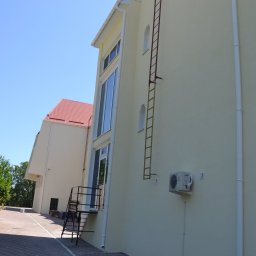 Denys Denysov - Elewacja budynku z zamontowaną drabiną ewakuacyjną i klimatyzatorem, widoczny fragment chodnika z kostki brukowej oraz budynek z czerwonym dachem w tle.