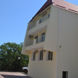 Denys Denysov - Trzypiętrowy budynek z beżową elewacją, balkonami z metalowymi barierkami i czerwonym dachem, widoczny z boku, z drzewami w tle i częściowo widocznym białym samochodem.