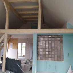 Denys Denysov - Poddasze w trakcie adaptacji: widoczne drewniane belki konstrukcyjne, ściany z płyt gipsowo-kartonowych w kolorze turkusowym, okno z luksferów oraz narzędzia i materiały budowlane na podłodze.