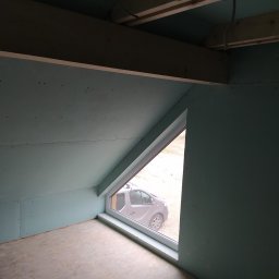 Denys Denysov - Poddasze z oknem w kształcie trapezu, ściany wykończone płytami gipsowo-kartonowymi, widoczne drewniane belki stropowe i instalacja elektryczna, na zewnątrz zaparkowany samochód.