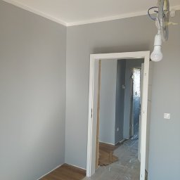 Denys Denysov - Świeżo pomalowane na szaro wnętrze z drewnianą podłogą, białą listwą przypodłogową i futryną, tymczasowe oświetlenie żarówką na kablu, podłoga zabezpieczona folią malarską.