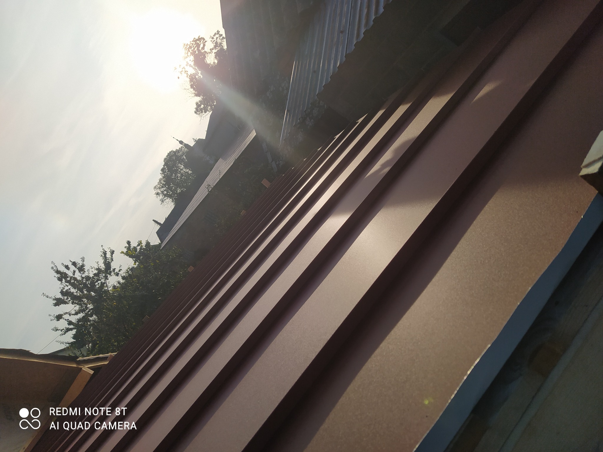 Nowe, brązowe blaszane pokrycie dachowe w promieniach słońca, widoczne drzewa i fragmenty budynków w tle.