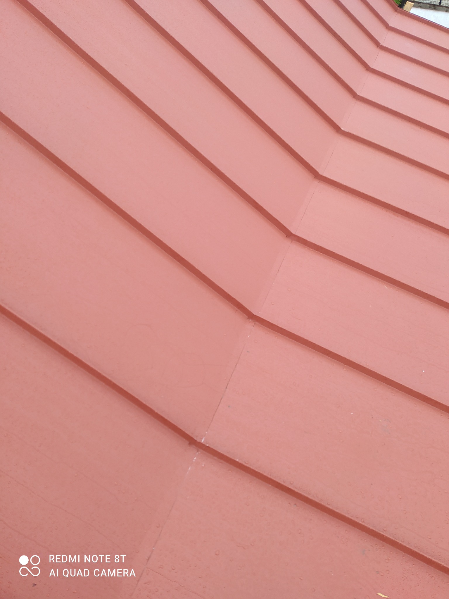 Czerwone metalowe panele dachowe ułożone ukośnie, widoczne łączenia i krople wody.