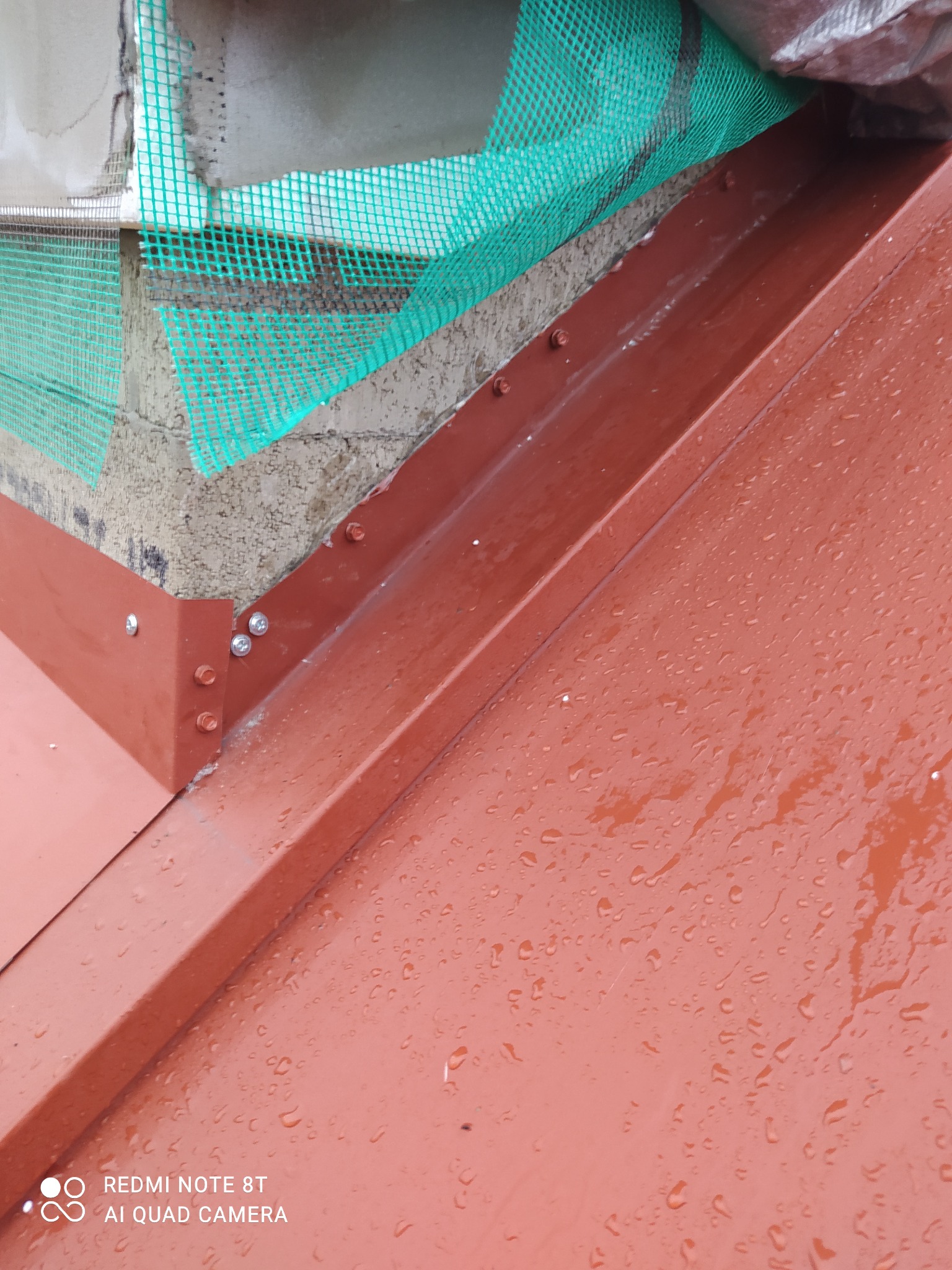 Czerwona blacha na dachu z widocznymi kroplami deszczu, mocowana na śruby, z fragmentem ściany zabezpieczonej zieloną siatką w tle.