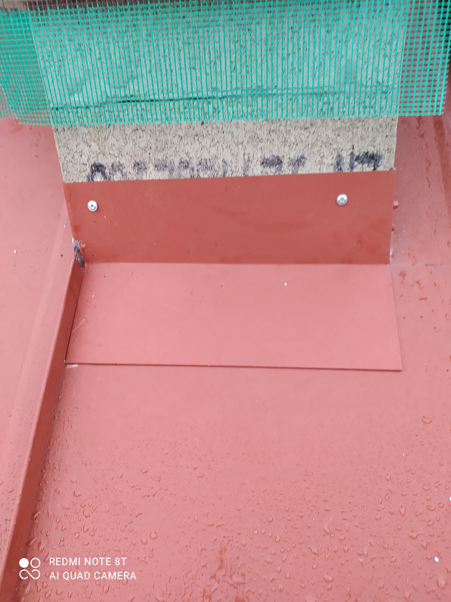 Szczegółowe ujęcie okapu dachu z blachy w kolorze ceglastym, z widocznymi śladami deszczu, mocowanego na wkręty do podłoża z siatką wentylacyjną.