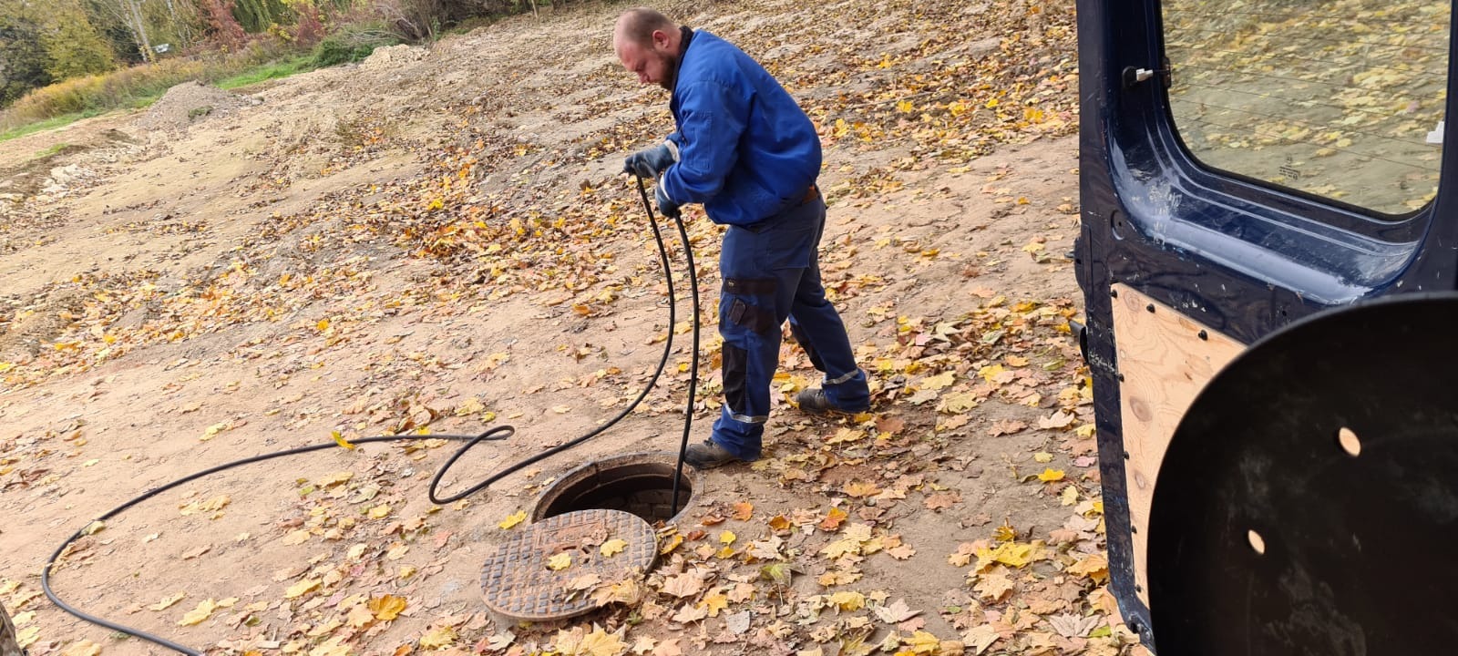 Hydraulik w niebieskim kombinezonie i rękawicach wprowadza wąż do czyszczenia kanalizacji do otwartego włazu studzienki na tle ziemi pokrytej jesiennymi liśćmi, obok otwarta boczna ściana samochodu...