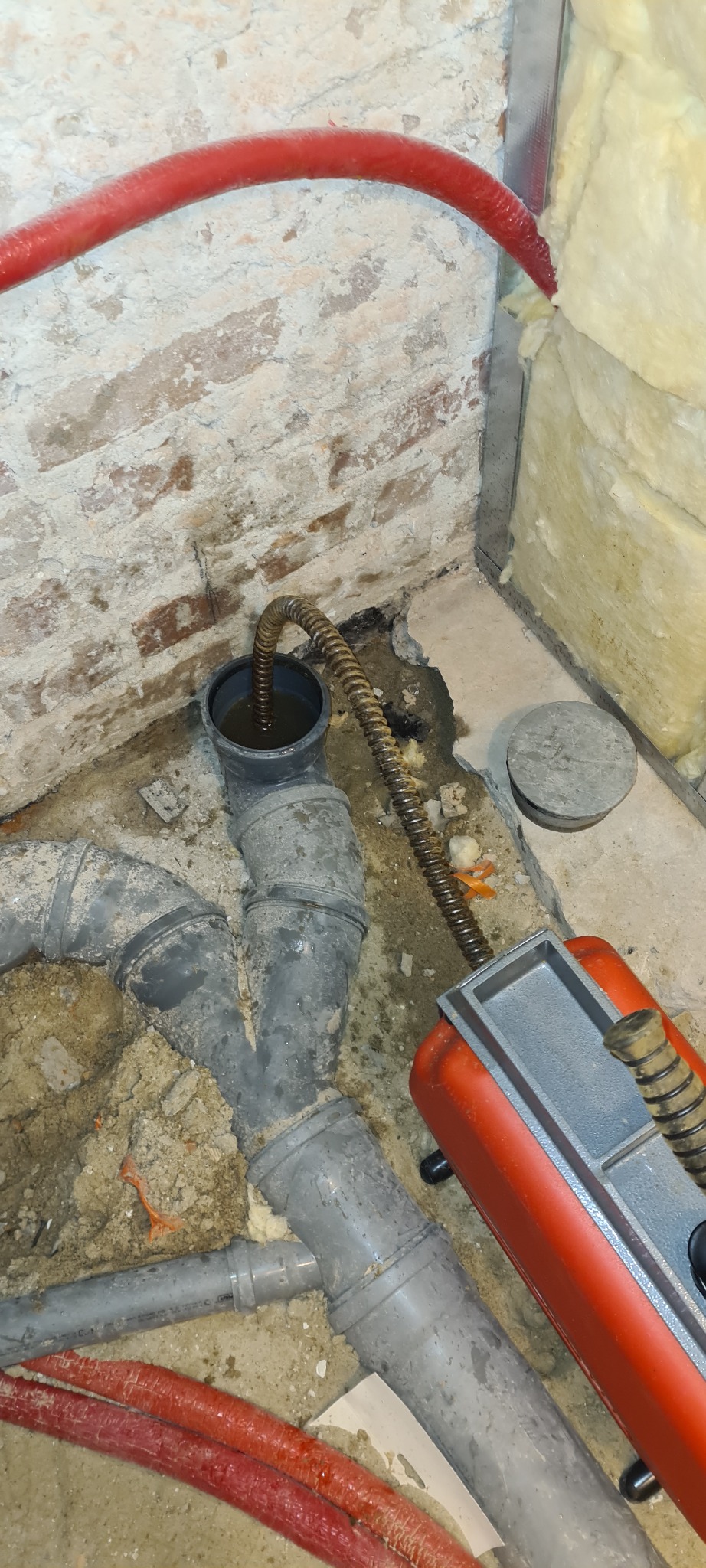 Spiralne narzędzie do czyszczenia wprowadzone do rury kanalizacyjnej wypełnionej wodą, widoczne szare rury PVC, czerwone kable i fragment ściany z cegły w tle.