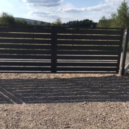 IG-NAT Ogrodzenia i bramy Grzegorz Błaszczyk - Nowoczesna, pozioma brama wjazdowa z metalowymi przęsłami w kolorze antracytowym, na tle błękitnego nieba z chmurami i zieleni drzew. Widok z zewnątrz posesji.