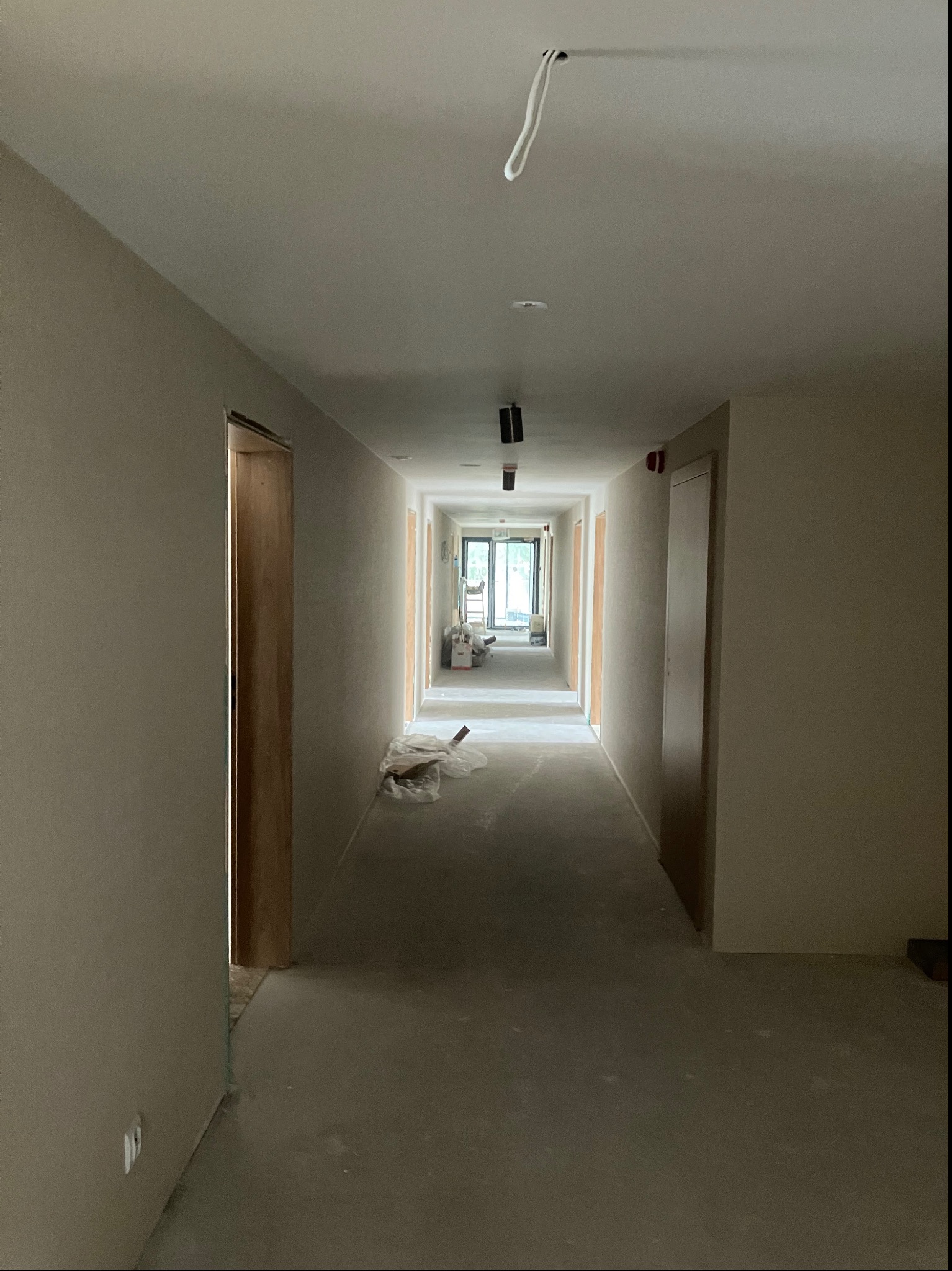 Długi korytarz w trakcie remontu, ściany przygotowane pod malowanie lub tapetowanie, widoczne ślady szpachlowania i zabezpieczenia podłogi, surowy stan wykończenia.