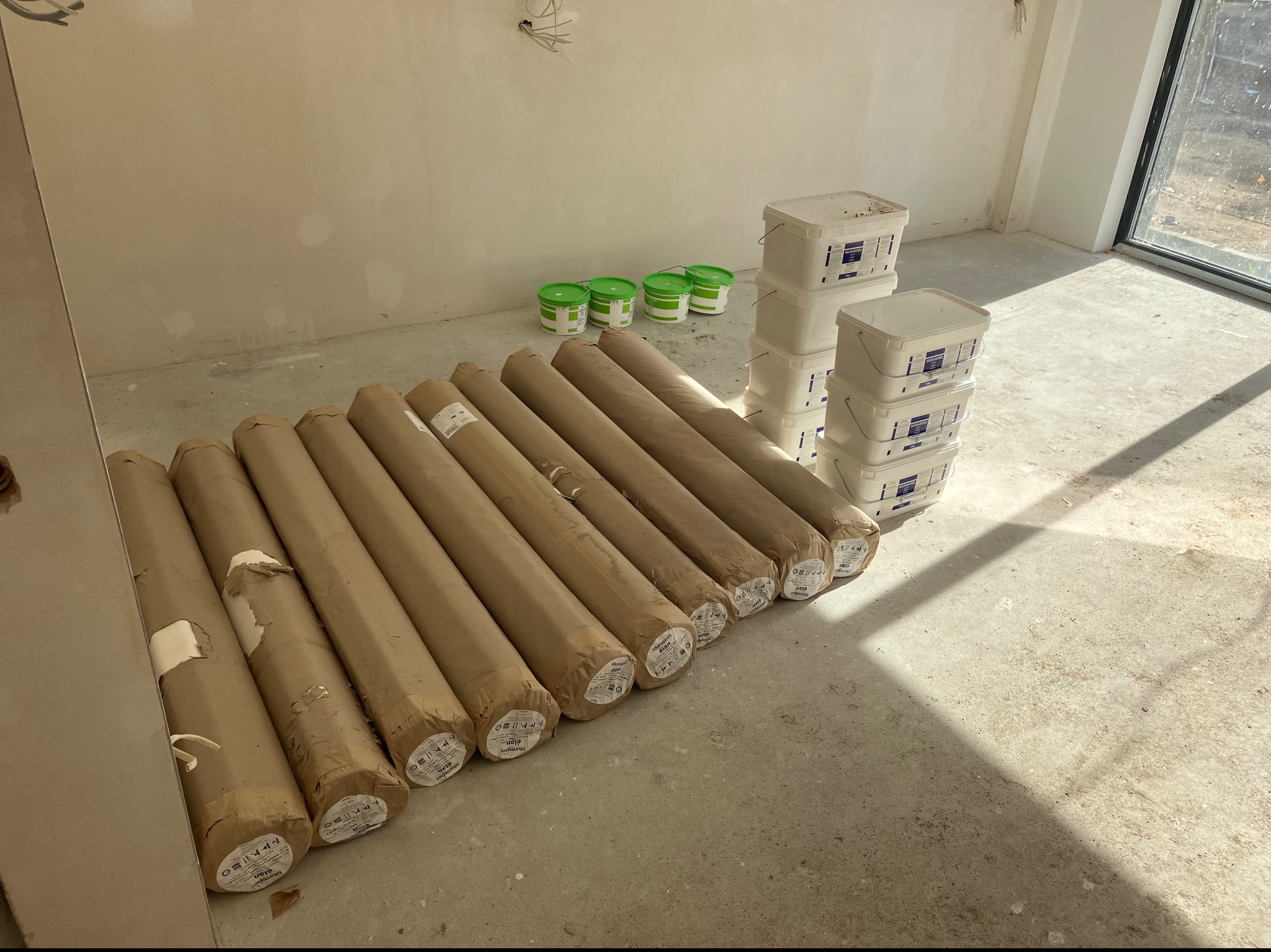 Przygotowane do montażu rolki tapet w brązowym papierze, białe wiadra z klejem i zielone wiadra z gruntem w jasnym, surowym pomieszczeniu z dużym oknem.