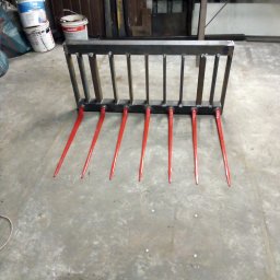 Goł-spaw - Konstrukcja metalowa z czerwonymi prętami, prawdopodobnie widły do ładowacza czołowego, w warsztacie z widocznymi farbami i elementami metalowymi w tle.