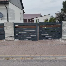 Nowoczesna brama wjazdowa z szarymi przęsłami i drewnianymi akcentami, osadzona na betonowych słupkach z bloczków, prowadząca na brukowany podjazd przed domem.