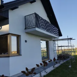 Nowoczesny dom z balkonem zabezpieczonym czarną, metalową balustradą, widoczne rusztowanie przy ścianie.