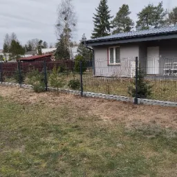 Nowoczesne ogrodzenie panelowe z podmurówką betonową, widok z zewnątrz na posesję z młodymi drzewkami iglastymi wzdłuż ogrodzenia i domem w tle.