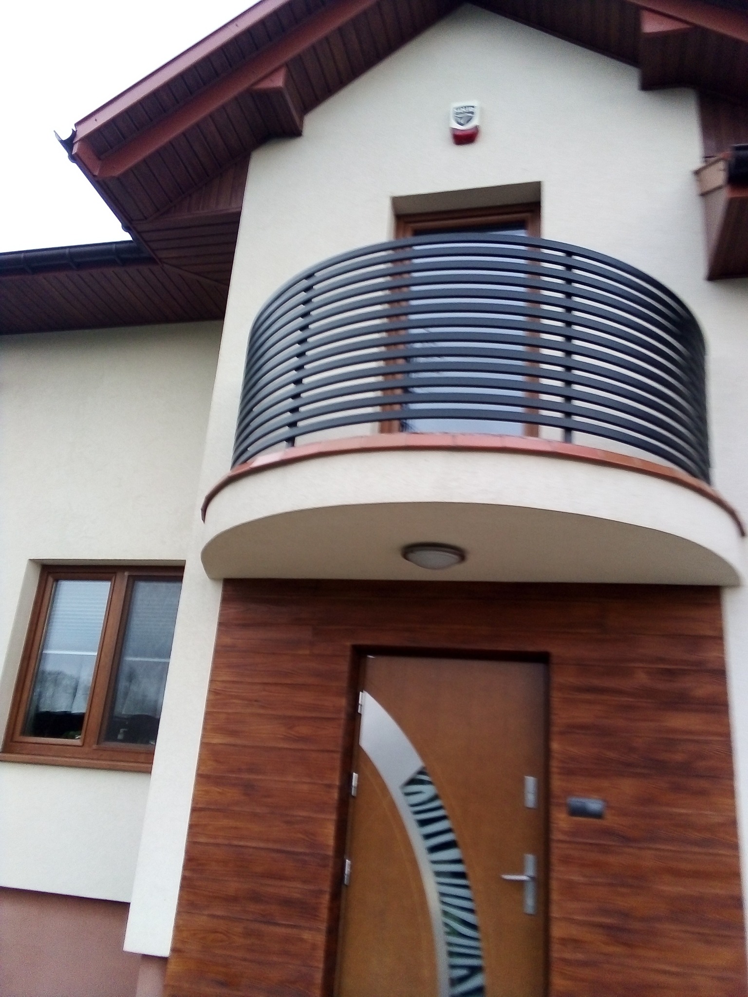Elewacja domu z zaokrąglonym balkonem z czarną, poziomą balustradą, drewnopodobną stolarką okienną i drzwiami wejściowymi z dekoracyjnym, srebrnym elementem.