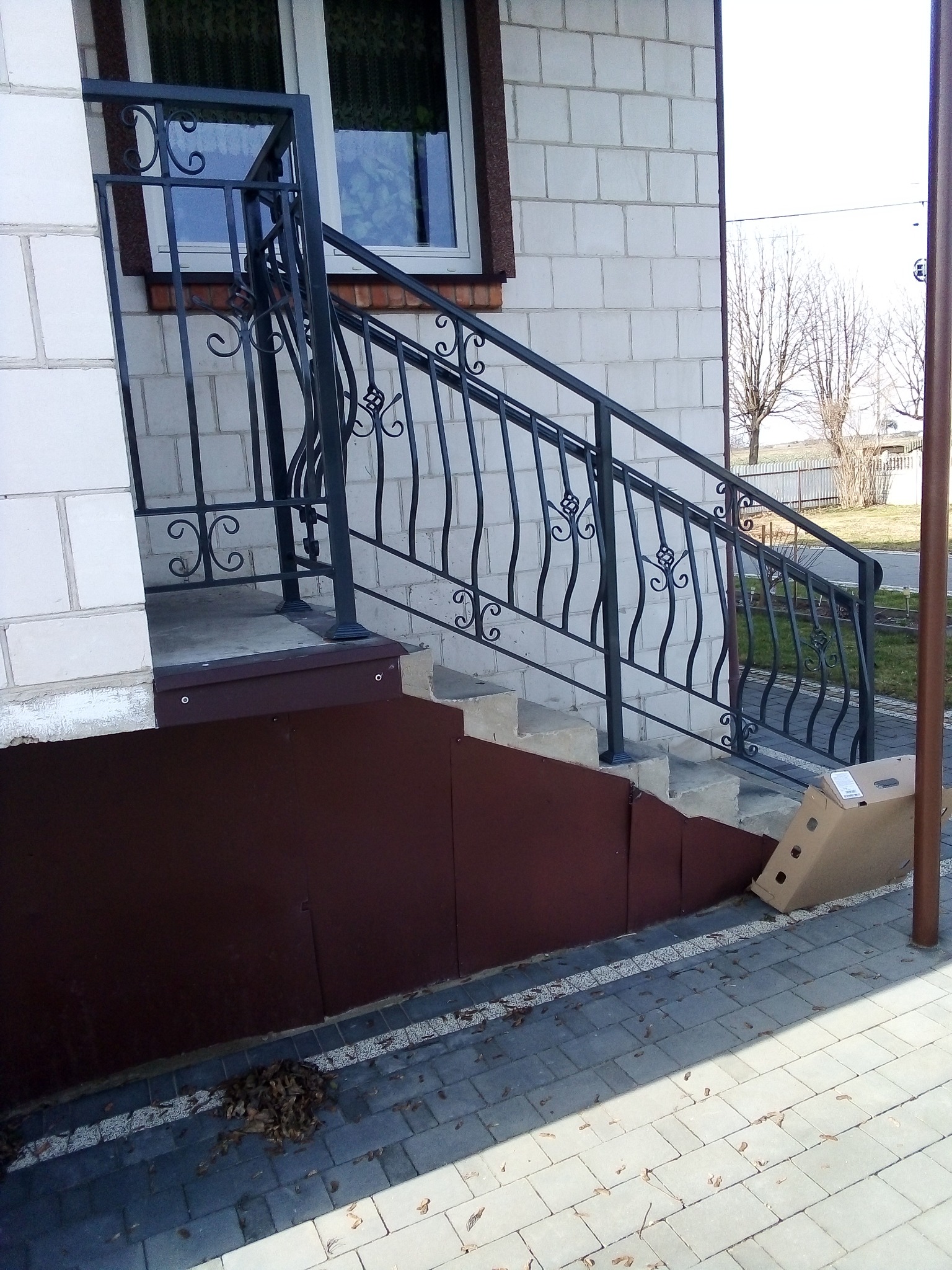 Czarne, metalowe balustrady z dekoracyjnymi elementami na schodach wejściowych do domu z białej cegły, częściowo zasłonięte kartonem, widok z dołu.