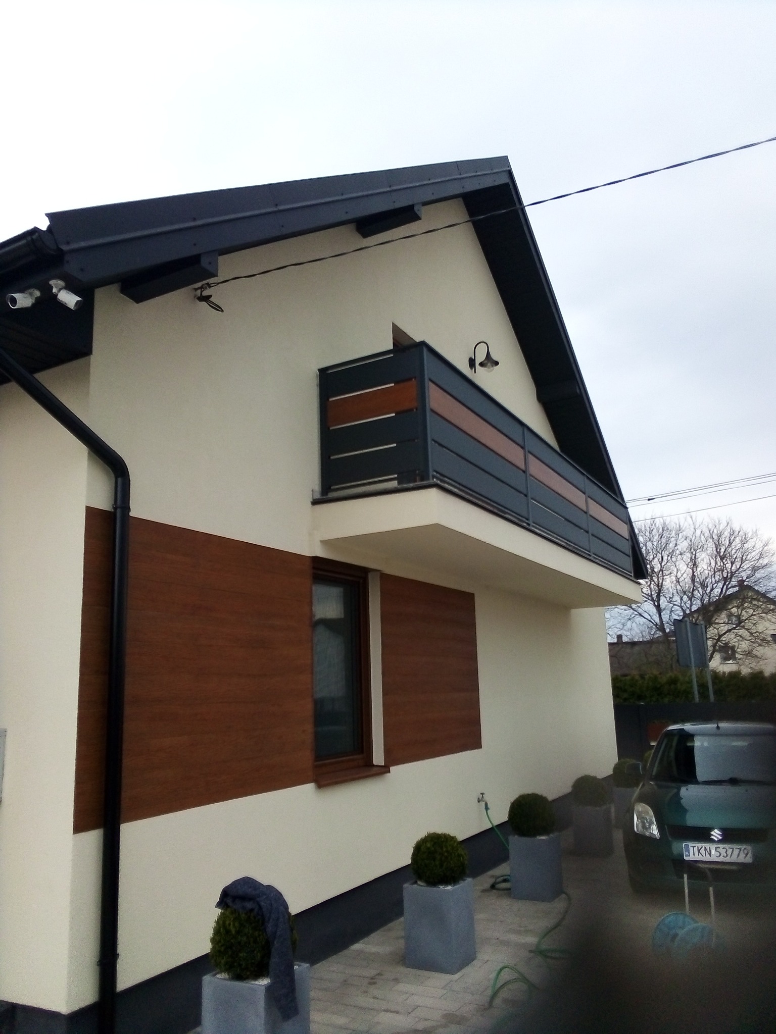 Elewacja domu z balkonem z nowoczesną balustradą, okładziną imitującą drewno i kamerami monitoringu, widok z zewnątrz.