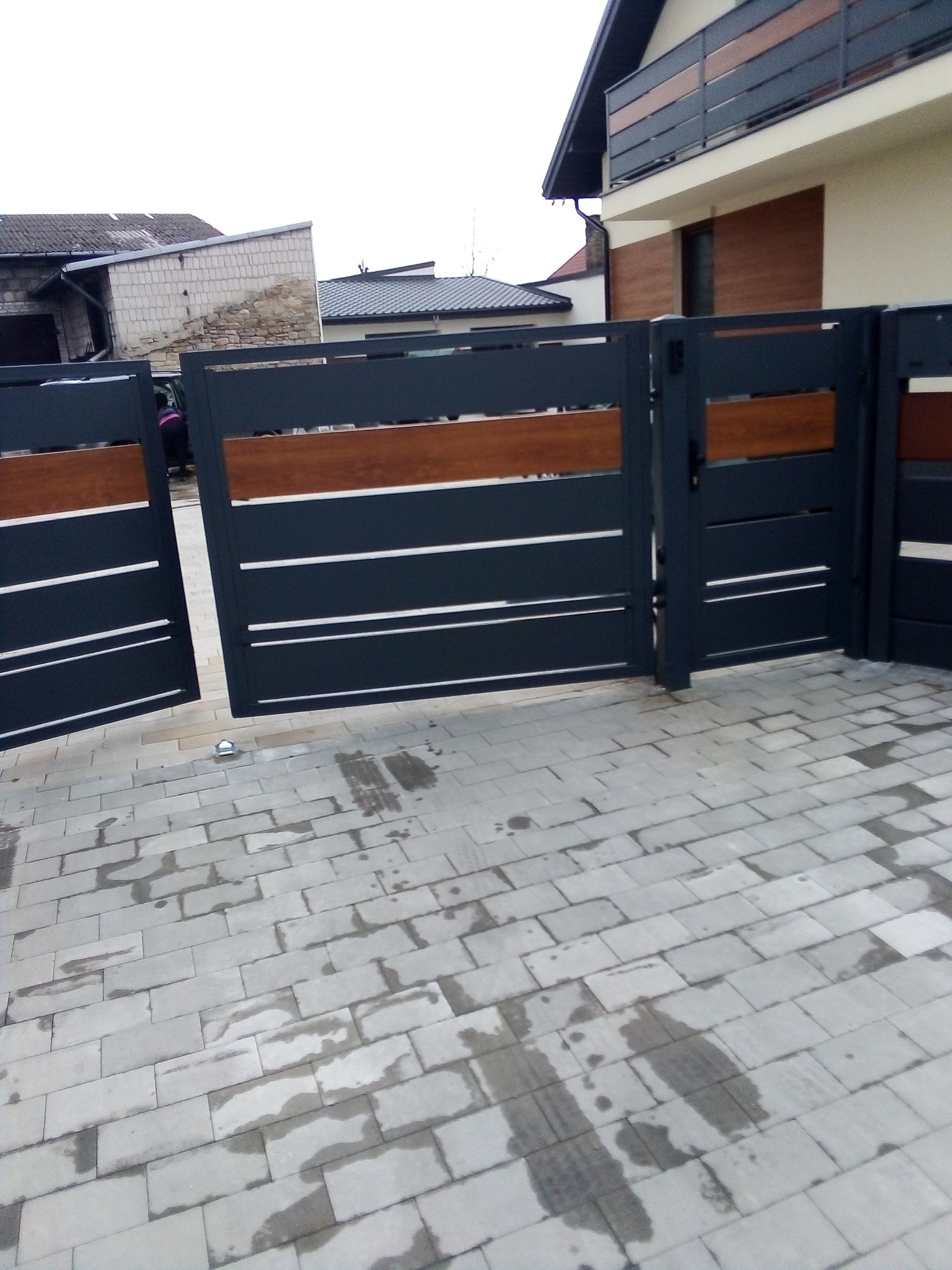 Nowoczesna, grafitowa brama wjazdowa z poziomymi, drewnianymi akcentami, otwierana na posesję wyłożoną szarą kostką brukową w pochmurny dzień.