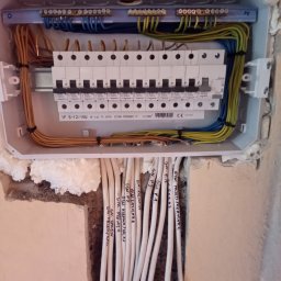 Elkom - Rozdzielnica elektryczna z zainstalowanymi bezpiecznikami Legrand, widoczne kable oznaczone opisami pomieszczeń, zamontowana w ścianie z pianką montażową.