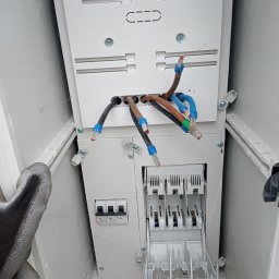 Elkom - Otwarta skrzynka elektryczna z wystającymi przewodami zakończonymi tulejkami, widoczne bezpieczniki i fragment dłoni w roboczej rękawicy.