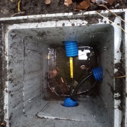 Elkom - Studzienka rewizyjna z niebieskimi rurami karbowanymi i czarnym kablem wewnątrz, wypełniona wodą i liśćmi, widok z góry.