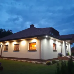 Dom jednorodzinny z brązowym dachem oświetlony punktowymi lampami LED pod okapem, widoczne okna z roletami i zadbany trawnik.