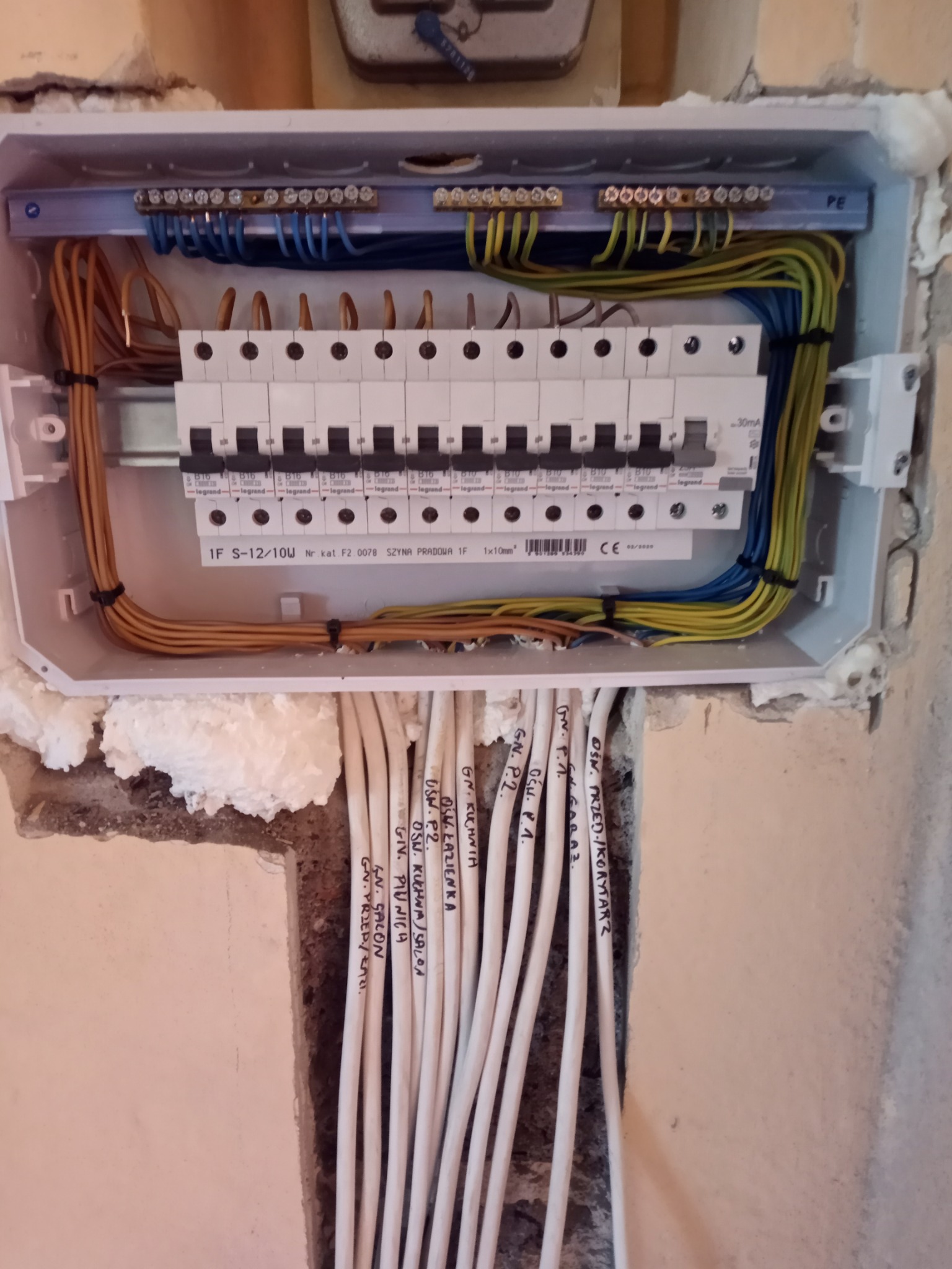 Rozdzielnica elektryczna z zainstalowanymi bezpiecznikami Legrand, widoczne kable oznaczone opisami pomieszczeń, zamontowana w ścianie z pianką montażową.