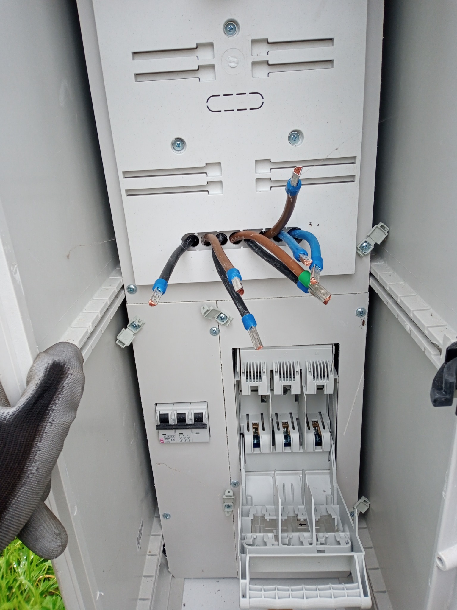 Otwarta skrzynka elektryczna z wystającymi przewodami zakończonymi tulejkami, widoczne bezpieczniki i fragment dłoni w roboczej rękawicy.