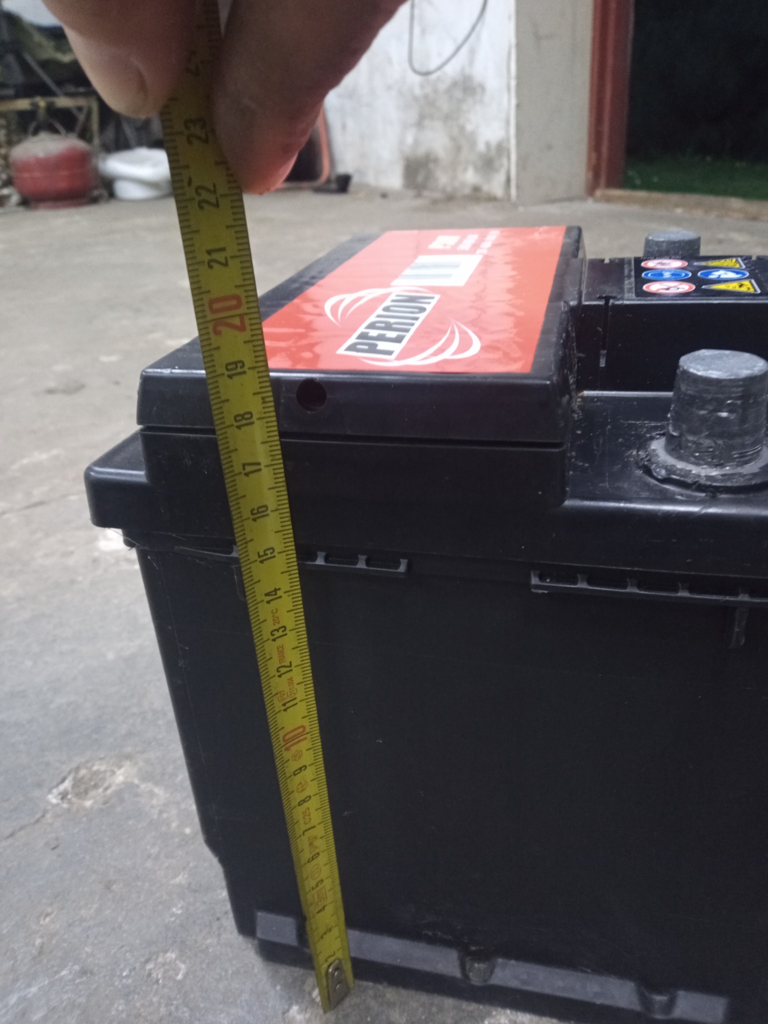 Pomiar wysokości akumulatora samochodowego marki Perion za pomocą żółtej miarki zwijanej, widoczne oznaczenia na akumulatorze i miarce.