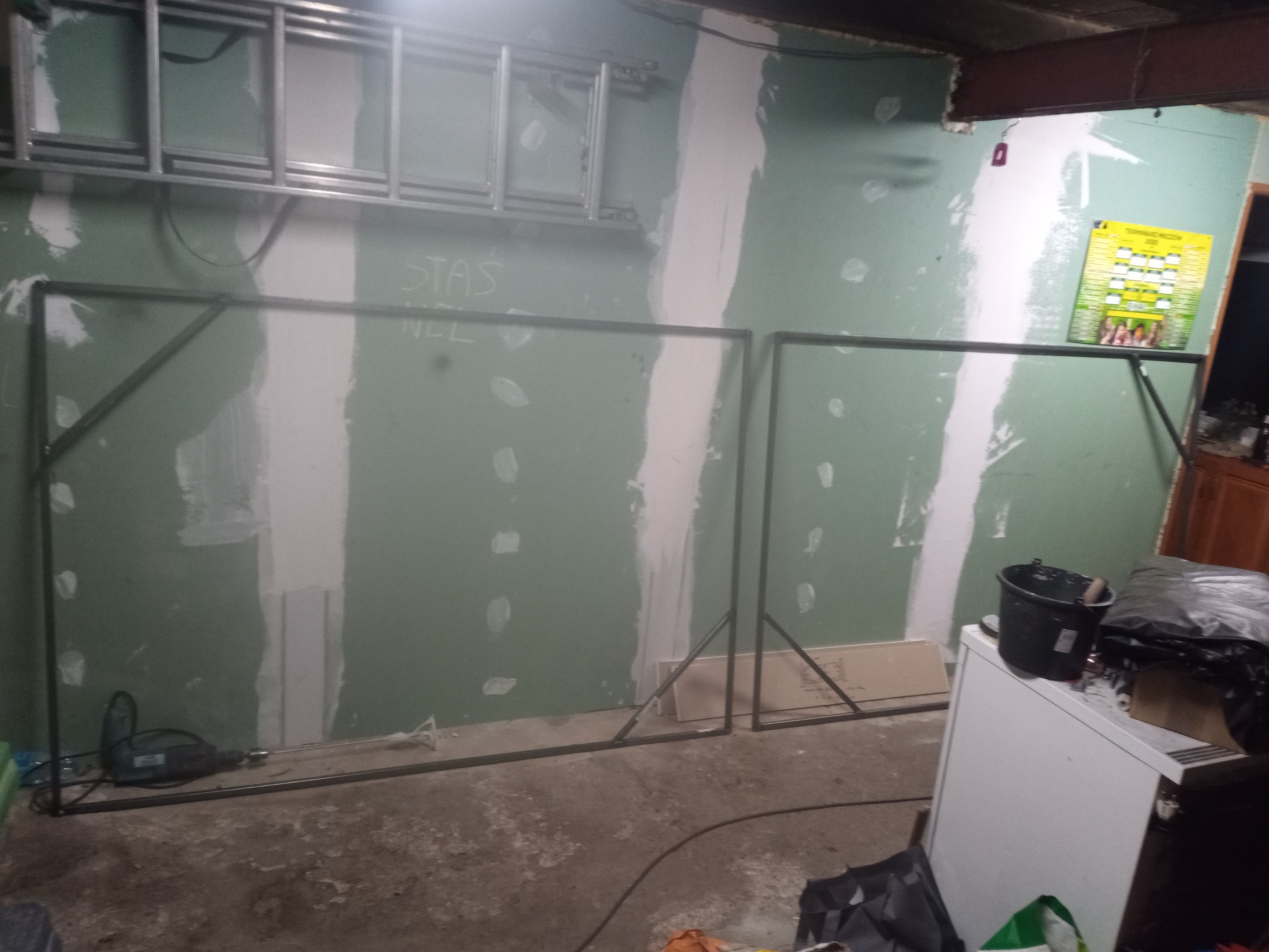 Metalowe ramy konstrukcyjne ustawione w surowym wnętrzu z częściowo wykończonymi ścianami gipsowymi, widoczna drabina na ścianie i narzędzia budowlane w tle.