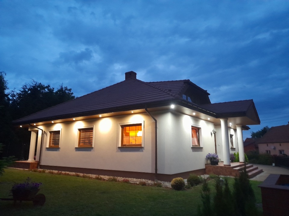 Dom jednorodzinny z brązowym dachem oświetlony punktowymi lampami LED pod okapem, widoczne okna z roletami i zadbany trawnik.