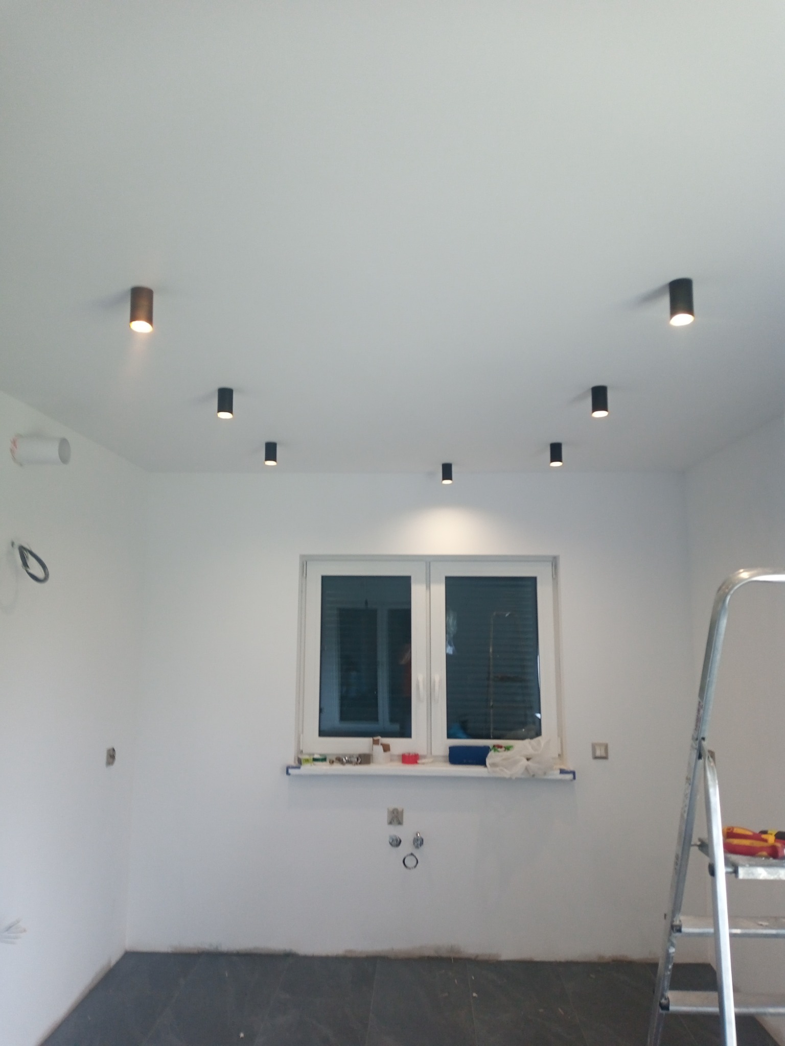 Zamontowane punktowe oświetlenie sufitowe w nowym pomieszczeniu, widoczna drabina i przygotowane gniazdka elektryczne.