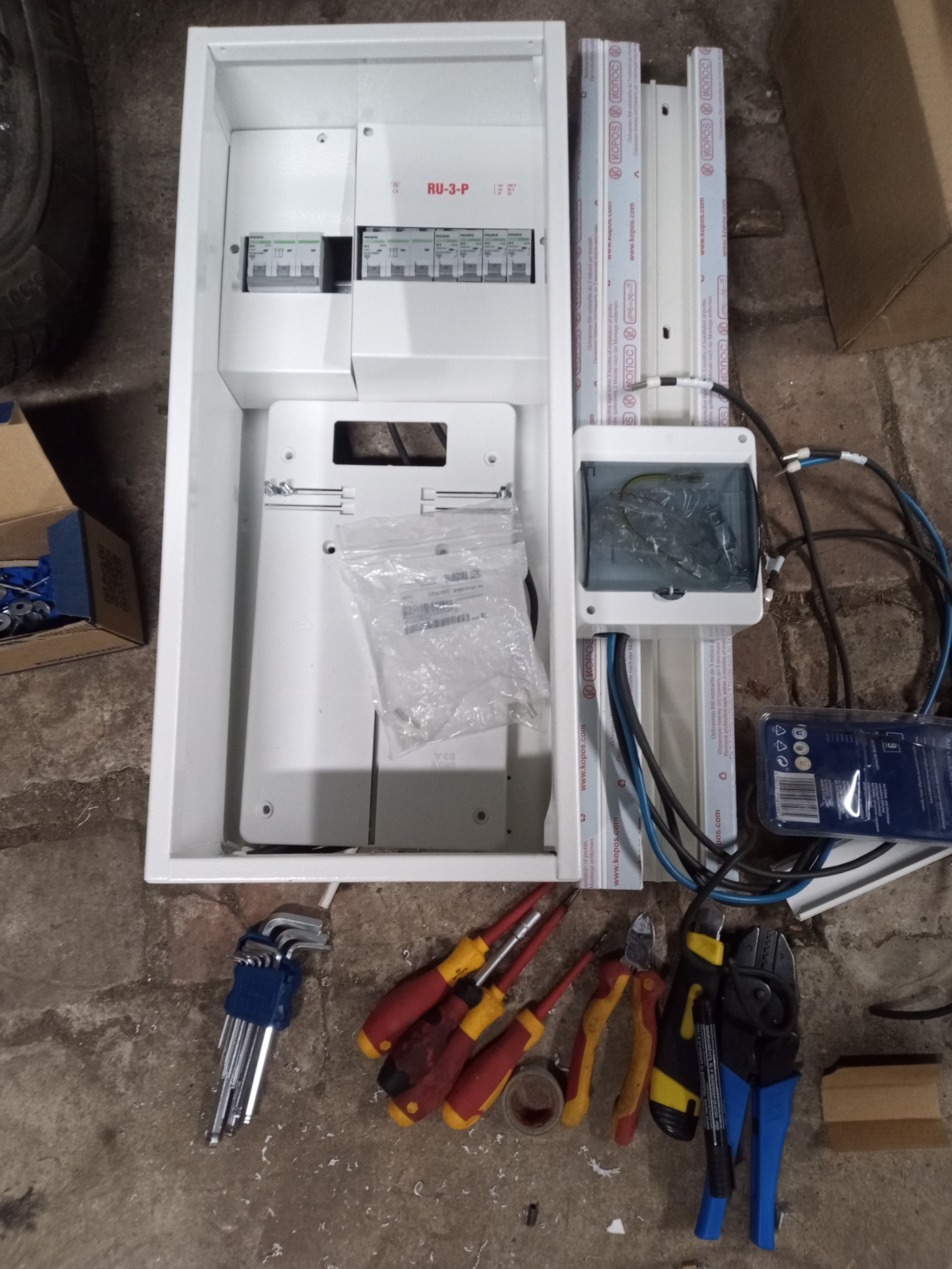 Rozdzielnica elektryczna w obudowie, wraz z narzędziami: śrubokręty, kombinerki, klucze imbusowe, obok puszka instalacyjna, listwy montażowe i przewody na szarym podłożu.